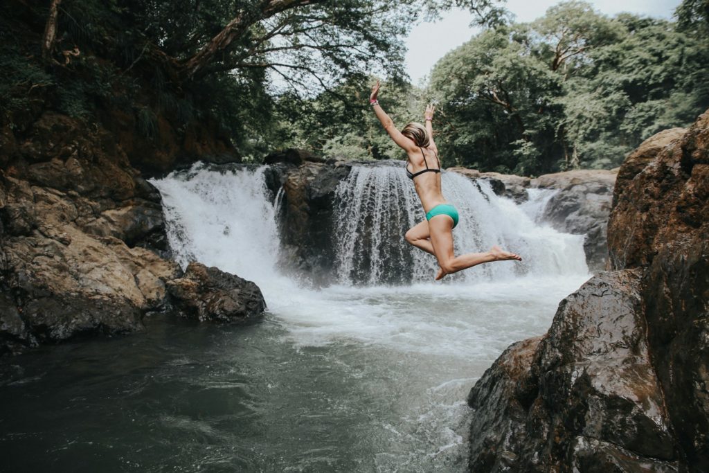 woman in bikini jumping off rocks into waterfall pool